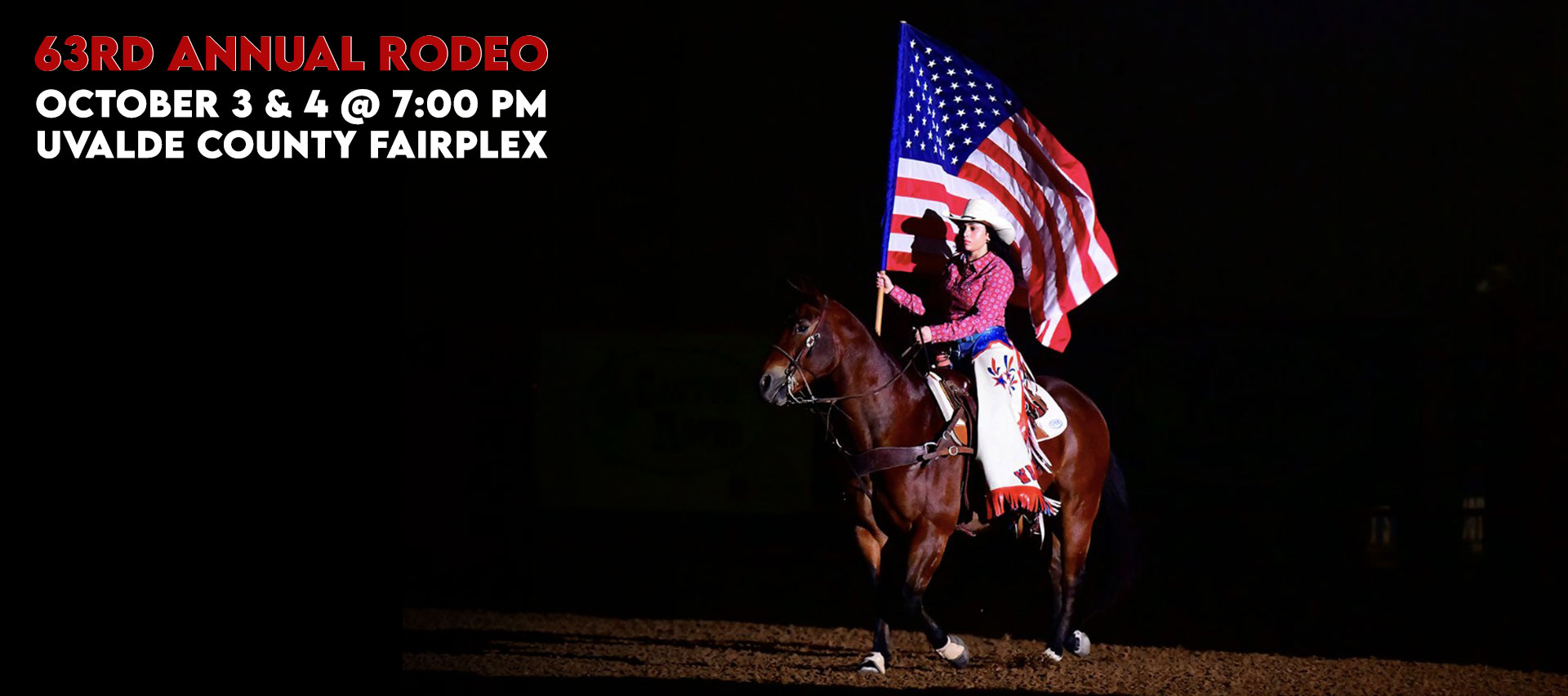 A rodeo rider in a cowboy hat and red shirt carries a large American flag while riding a brown horse under a spotlight in a dark arena.
