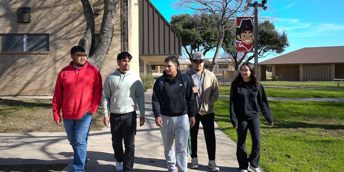 Five students walking on a sidewalk near the Uvalde Campus gym.