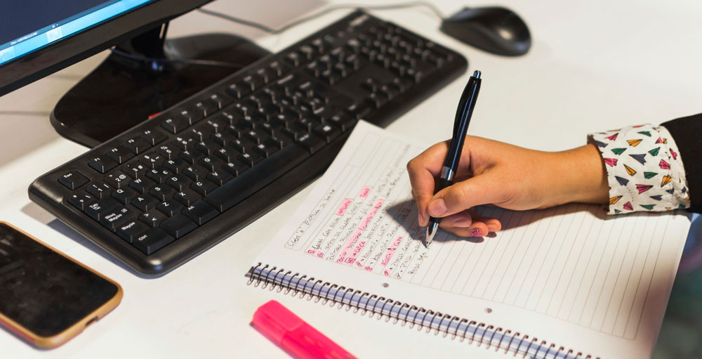 Picture of a person writing notes on a notepad in front of a computer.